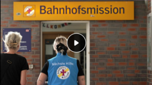 Eine Mitarbeiterin der Bahnhofsmission in blauer Weste und eine Frau betreten die Bahnhofsmission in Münster (Titelbild des Videos)